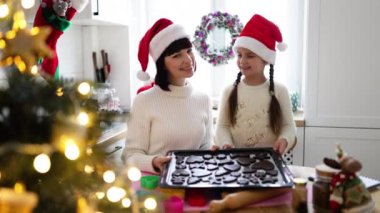 Gülümseyen genç bir kadın ve kız Noel Baba şapkası takıp sıcak mutfakta kurabiye tepsisine kurabiye koyuyorlar. Sahne, bayram ruhunu, aile bağlarını ve şenlik aktivitelerini yakalıyor.