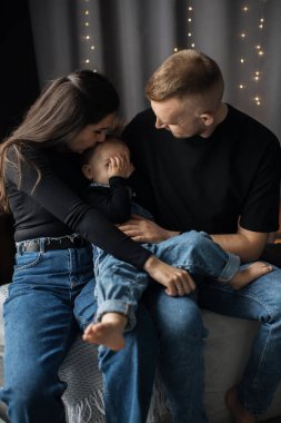 Genç Kafkasyalı anne ve baba kanepede oğluyla birlikte oturuyor, sevgi dolu bir anı paylaşıyorlar. Anne çocuğun alnını öperken, baba sevgiyle izliyor..