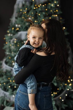 Genç beyaz anne, güzel dekore edilmiş bir Noel ağacının önünde, sıcaklığı ve mutluluğu yakalayarak kızını neşeyle kucaklıyor..