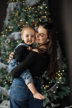 Genç beyaz anne, güzel dekore edilmiş bir Noel ağacının önünde, sıcaklığı ve mutluluğu yakalayarak kızını neşeyle kucaklıyor..