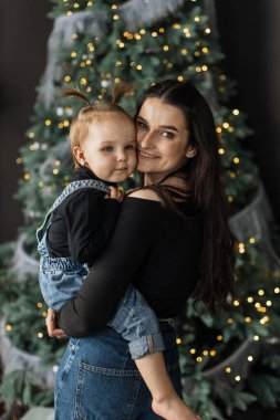 Genç beyaz anne, güzel dekore edilmiş bir Noel ağacının önünde, sıcaklığı ve mutluluğu yakalayarak kızını neşeyle kucaklıyor..