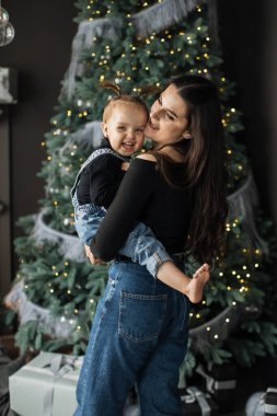 Genç beyaz anne, gülümseyen bebeği süslü Noel ağacının yanında tutuyor. İkisi de kot giyiyor, sıcak, sevgi ve tatil ruhu taşıyor. Manzara neşe ve şenlikli atmosferle dolu..