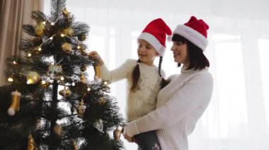 Gülümseyen beyaz anne ve kız Noel Baba şapkaları takarak Noel ağacını altın süslemeler ve ışıklarla süslüyorlar. Tatil döneminde evde şenlikli atmosfer, mutluluk ve aile bağları.