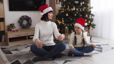 Kafkasyalı anne ve kız yoga pozisyonlarında oturuyor, Noel Baba şapkaları takıyor, Noel ağacıyla süslenmiş sıcak oturma odasında birlikte meditasyon yapıyorlar..