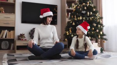 Beyaz anne ve kız, Noel ağacının yanında, Noel Baba şapkası takarak yoga pozisyonunda oturuyorlar. Sıcak tatil atmosferi, bayram sezonunda rahatlama ve farkındalık ifade eder..