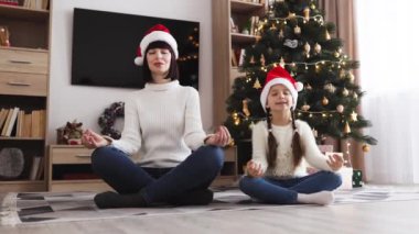Kafkas anne ve kızı yoga pozisyonunda oturmuş Noel Baba şapkaları takıyor Noel ağacının yanında meditasyon yapıyor ve huzurlu bir tatil ortamı yaratıyor. Işıklarla süslenmiş ağaç ısınıyor..