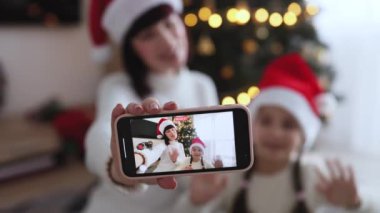 Beyaz kadın ve Noel Baba şapkası takan genç kız cep telefonuyla video görüşmesi yapıyor. Noel ağacının önünde oturmuş, neşe ve tatil ruhunu ifade ediyor..