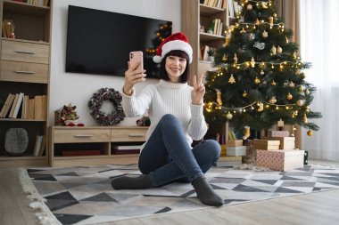 Beyaz kadın Noel Baba şapkası takıyor ve akıllı telefondan video görüşmesi yapıyor. Süslü Noel ağacı ve hediyelerle dolu bir oturma odasında..