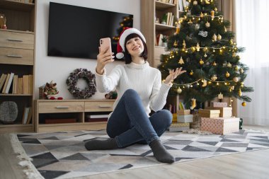 Noel Baba şapkası takan neşeli beyaz kadın akıllı telefondan video görüşmesi yapıyor. Süslü Noel ağacının yanında parlak ışıklar ve rahat oturma odasında hediyelerle yan yana oturuyor..