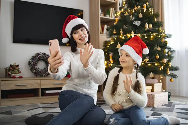 Noel Baba şapkası takan beyaz anne ve kız Noel ağacının önünde yerde otururken cep telefonlarıyla video görüşmesi yapıyorlar. Tatil sezonunun ve evde neşeli bir atmosferin tadını çıkarıyorum..