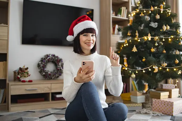 Noel Baba şapkalı genç beyaz kadın elinde akıllı telefon Noel ağacının yanında oturuyor. Gülümsüyor ve yukarı doğru işaret ediyor, sıcak oturma odasında neşe ve tatil ruhunu ifade ediyor..