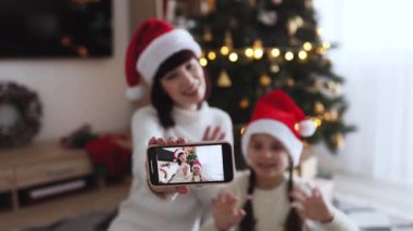 Beyaz kadın ve Noel Baba şapkası takan genç kız cep telefonuyla video görüşmesi yapıyor. Noel ağacının önünde oturmuş, neşe ve tatil ruhunu ifade ediyor..