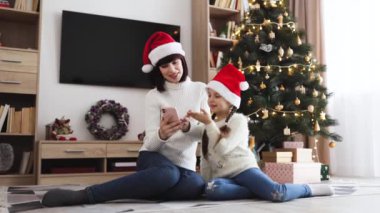 Beyaz anne ve kız Noel ağacının önünde yerde telefon kullanırken Noel Baba şapkası takıyor. Tatil sezonunda çekilen neşeli aile anı, bağlantıyı ve geleneği ön plana çıkarıyor.