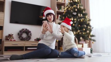 Kafkasyalı anne ve kız Noel Baba şapkası takıyor. Cep telefonuyla kutlama videosu çekiyor. Güzel dekore edilmiş Noel ağacının yanında otururken..