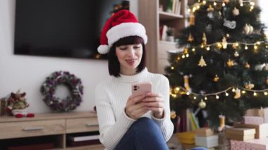 Noel 'de ağaç ve çelenkle süslenmiş rahat oturma odasında Noel Baba şapkası takan gülümseyen beyaz kadın. Tatil ruhunu ve neşesini yakalar, teknoloji ve geleneği harmanlar.