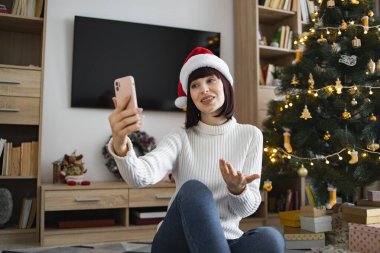 Noel Baba şapkası takan neşeli beyaz kadın akıllı telefondan video görüşmesi yapıyor. Noel ağacının yanında oturmak, evde tatil sezonunun tadını çıkarmak..