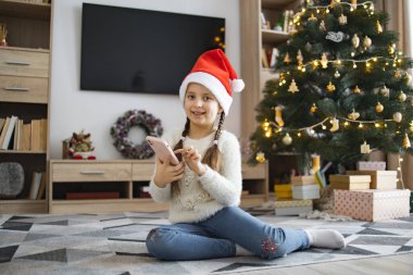 Noel Baba şapkalı genç beyaz kız Noel boyunca akıllı telefon kullanarak yerde oturuyor. Arkaplanda süslenmiş ağaç ve paketlenmiş hediyeler yer alıyor ve bayram atmosferi yaratılıyor.