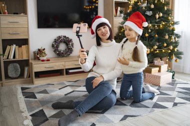 Kafkasyalı anne ve kız Noel şapkalı. Video görüşmesi veya selfie için akıllı telefon kullanıyorlar. Oturma odasında oturup gülümsüyorlar ve birlikte tatil sezonunun tadını çıkarıyorlar..