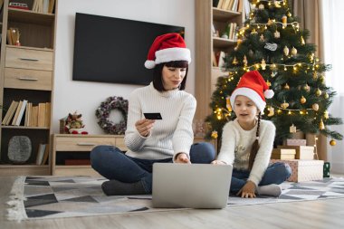 Noel Baba şapkası takan beyaz kadın ve genç kız Noel tatilinde kredi kartıyla online alışveriş yapıyorlar. Oturma odasının zemininde, dekore edilmiş ağacın yanında..