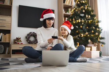 Kafkasyalı anne ve kız Noel Baba şapkası takıyor ve bilgisayar ve kredi kartıyla online alışveriş yapıyorlar. Noel ağacı süslenmiş ışıklar ve hediyeler rahat oturma odası düzeninde..