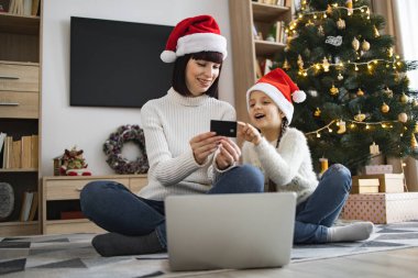 Noel Baba şapkası takan beyaz anne ve kız banka kartı kullanarak Noel indirimleri sırasında online alışveriş yapıyorlar. Süslü ağacın yanında otururken, her ikisi de bayram ruhunun keyfini çıkarıyor..