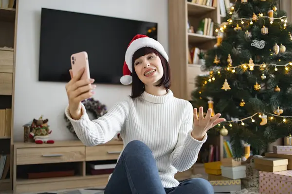 Beyaz kadın Noel Baba şapkası takıyor ve akıllı telefondan video görüşmesi yapıyor. Süslü Noel ağacı ve hediyelerle sıcacık ev ortamında gülümsüyor. Bayram sezonunda şenlik ruhunu yakalar.