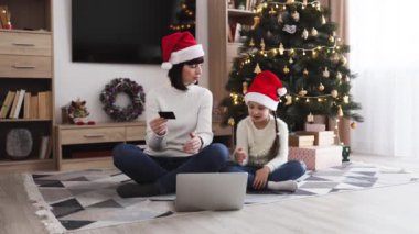 Kafkasyalı anne ve kız Noel Baba şapkası takıyor, Noel indirimleri sırasında banka kartı kullanarak internetten alışveriş yapıyor. Noel ağacı ve dizüstü bilgisayarla birlikte oturma odasında oturuyorlar..