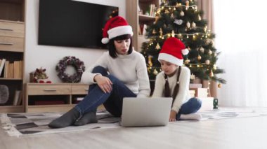 Noel Baba şapkası takan beyaz anne ve kız dizüstü bilgisayar kullanarak yerde oturuyorlar. Arka planda süslemeleri olan Noel ağacı şenlikli atmosfer yaratır.