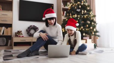 Noel Baba şapkası takan beyaz anne ve kız dizüstü bilgisayar kullanarak yerde oturuyorlar. Arka planda süslemeleri olan Noel ağacı şenlikli atmosfer yaratır.