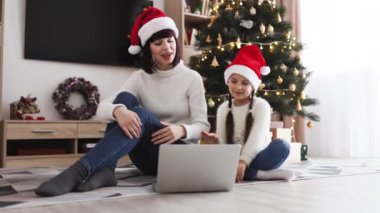 Noel Baba şapkası takan beyaz bir anne ve kız, Noel 'de evde online video görüşmesi için dizüstü bilgisayar kullanıyor. Noel ağacı ve hediyelerle süslenmiş bir oda bayram atmosferi yaratır..