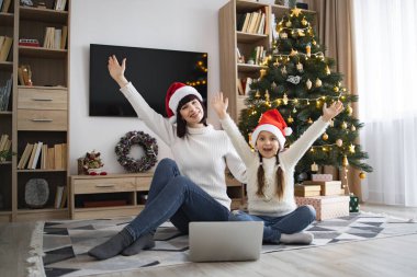 Mutlu Kafkasyalı anne ve kız Noel Baba şapkası takıp evde birlikte dizüstü bilgisayar kullanıyorlar. Gülümseyen aile festival sezonunun tadını çıkarıyor.