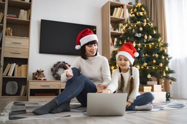 Kafkasyalı anne ve genç kız internette video görüşmesi için dizüstü bilgisayar kullanarak Noel Baba şapkası takıyor. Konforlu oturma odasındaki dekore edilmiş Noel ağacının yanında oturmuş birlikte tatil sezonunun tadını çıkarıyorduk..