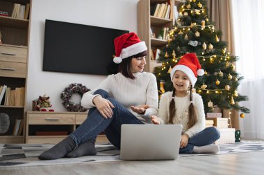 Kafkasyalı anne ve genç kız internette video görüşmesi için dizüstü bilgisayar kullanarak Noel Baba şapkası takıyor. Noel süsleri, ağaç ve hediyelerle çevrili..