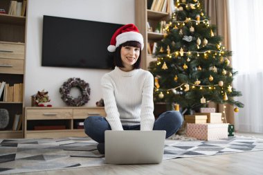 20 'li yaşlarda, Noel Baba şapkası takan, dizüstü bilgisayarlı, Noel ağacının yanında oturan beyaz bir kadın. Dekoratif ev dekorasyonu, hediyeler ve rahat bir ortam içerir..