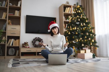 Genç beyaz kadın, video görüşmesi sırasında dizüstü bilgisayarla yerde oturan Noel Baba şapkası takıyor. Arka planda ışıklar ve süslerle süslenmiş Noel ağacı. Sıcak tatil atmosferi ve şenlik havası.