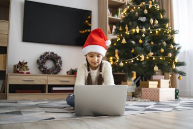 Beyaz kız Noel Baba şapkası takıyor ve Noel ağacının yanında dizüstü bilgisayar kullanıyor. Çocuk, şenlikli dekorasyonlar, hediyeler ve sıcak tatil atmosferiyle çevrili rahat oturma odasında yerde oturuyor..