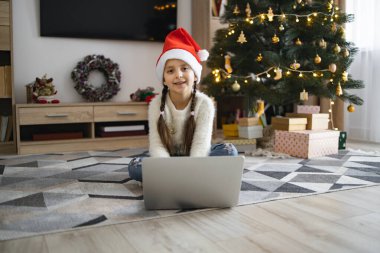 Kafkasyalı bir kız, 7 yaşlarında, Noel Baba şapkası takıyor, Noel ağacının yanında dizüstü bilgisayarla oturuyor. Şenlikli ışıklar ve hediyelerle süslenmiş bir oda, rahat bir tatil ortamı yaratıyor..