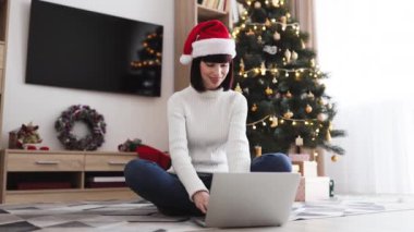 Noel Baba şapkası takan genç beyaz kadın, dekore edilmiş Noel ağacının yanında dizüstü bilgisayar kullanarak yerde oturuyor. Arkaplanda hediyeler ve çelenkler olan samimi bir ortam. Tatilleri evde kutluyoruz..