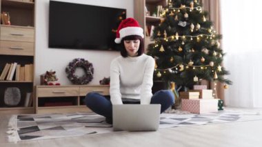 20 'li yaşlarda, Noel Baba şapkası takan, dizüstü bilgisayarlı, Noel ağacının yanında oturan beyaz bir kadın. Dekoratif ev dekorasyonu, hediyeler ve rahat bir ortam içerir..