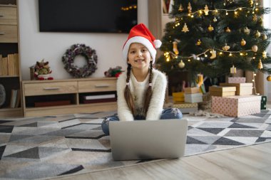 Noel Baba şapkalı genç beyaz kız Noel ağacının yanında dizüstü bilgisayar kullanarak yerde oturuyor. Gülümseyen çocuk bayram sezonunun tadını çıkarıyor, hediyeler ve dekorasyonla çevrili..