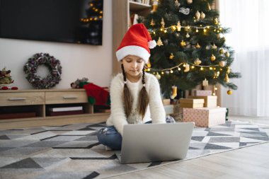 7-10 yaşlarında beyaz bir kız Noel Baba şapkası takıyor ve Noel ağacının yanında dizüstü bilgisayar kullanıyor. Konforlu oturma odasında, odaklanmış ve hediyelerle çevrili tatil ruhunun keyfini çıkarıyor..