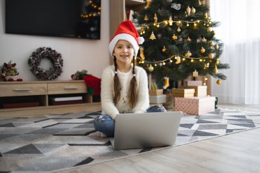 Noel Baba şapkası takan gülümseyen beyaz kız Noel ağacının yanında dizüstü bilgisayarla oturuyor. Etraftaki şenlikli dekorasyonlar neşeli bir tatil atmosferi olduğunu gösteriyor..