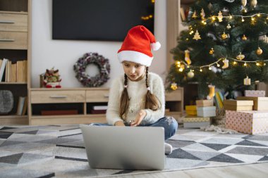 Noel Baba şapkası takan genç beyaz kız internette video görüşmesi yapıyor. Arka planda süslemeleri ve hediyeleri olan Noel ağacı şenlikli bir ortam yaratır..