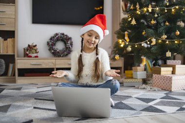 Çevrimiçi video görüşmesi sırasında dizüstü bilgisayar kullanarak yerde oturan ve Noel Baba şapkası takan gülümseyen genç kız. Arka planda ışıklar olan dekore edilmiş Noel ağacı. Hediyeler ve tatil dekorları şenlikli bir atmosfer yaratır..