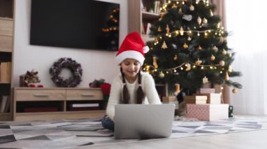 Kafkasyalı bir kız, 7 yaşlarında, Noel Baba şapkası takıyor, Noel ağacının yanında dizüstü bilgisayarla oturuyor. Şenlikli ışıklar ve hediyelerle süslenmiş bir oda, rahat bir tatil ortamı yaratıyor..