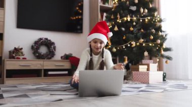 Noel Baba şapkası takan genç beyaz kız internette video görüşmesi yapıyor. Arka planda süslemeleri ve hediyeleri olan Noel ağacı şenlikli bir ortam yaratır..