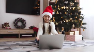 Çevrimiçi video görüşmesi sırasında dizüstü bilgisayar kullanarak yerde oturan ve Noel Baba şapkası takan gülümseyen genç kız. Arka planda ışıklar olan dekore edilmiş Noel ağacı. Hediyeler ve tatil dekorları şenlikli bir atmosfer yaratır..