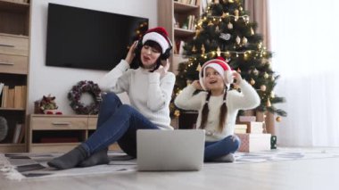 Noel ağacının arka planındaki şenlik sezonunda aile eğlencesi. Kafkasyalı genç bir anne ve küçük bir kız, dizüstü bilgisayar kullanarak kulaklıkla ilahiler söylerken,.