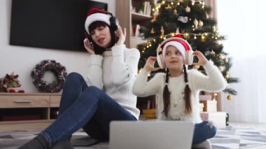 Noel ağacının arka planındaki şenlik sezonunda aile eğlencesi. Kafkasyalı genç bir anne ve küçük bir kız, dizüstü bilgisayar kullanarak kulaklıkla ilahiler söylerken,.