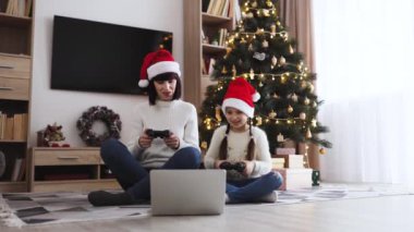 Noel Baba şapkası takan beyaz anne ve kız birlikte bilgisayar ve joystickle oyun oynuyorlar. Noel ağacı ve dekorasyonlar şenlikli bir aile ortamı yarattı.